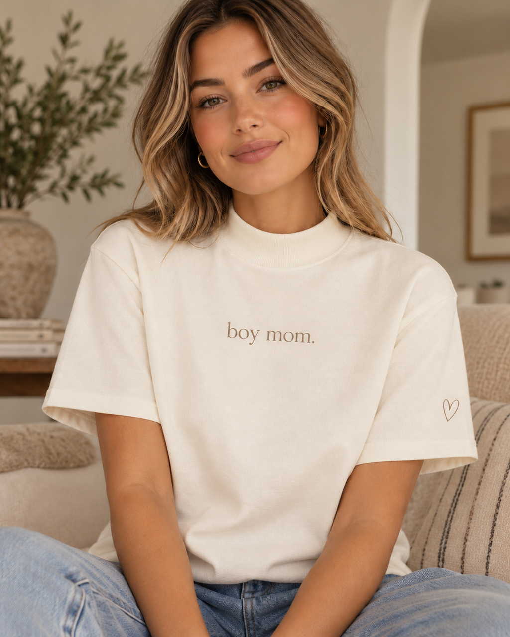 A woman wearing a cream-colored short-sleeve t-shirt with 'boy mom.' printed in brown text on the chest and a small brown heart outline on the sleeve, sitting on a couch in a living room.
