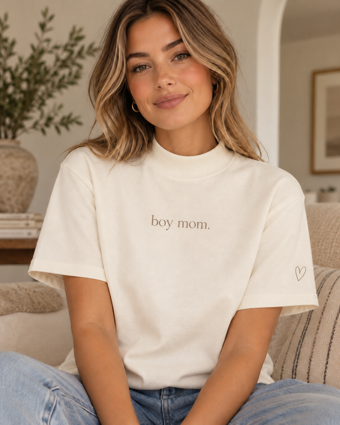 A woman wearing a cream-colored short-sleeve t-shirt with 'boy mom.' printed in brown text on the chest and a small brown heart outline on the sleeve, sitting on a couch in a living room.